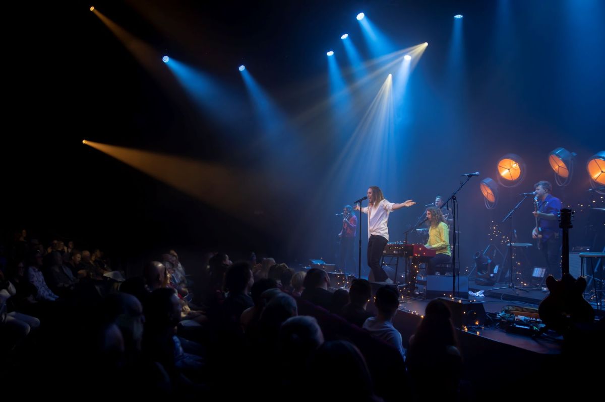 Tim Minchin and band performing ‘Tim Minchin – First at the Foundry’ at the Foundry Theatre, Sydney. The photo shows Minchin, a fair-skinned man with shoulder length hair and wearing a white shirt and dark trousers, caught mid-song. His arms are stretched wide and four musicians are visible on the stage behind him; the other half of the photo shows the audience members at the Foundry Theatre.