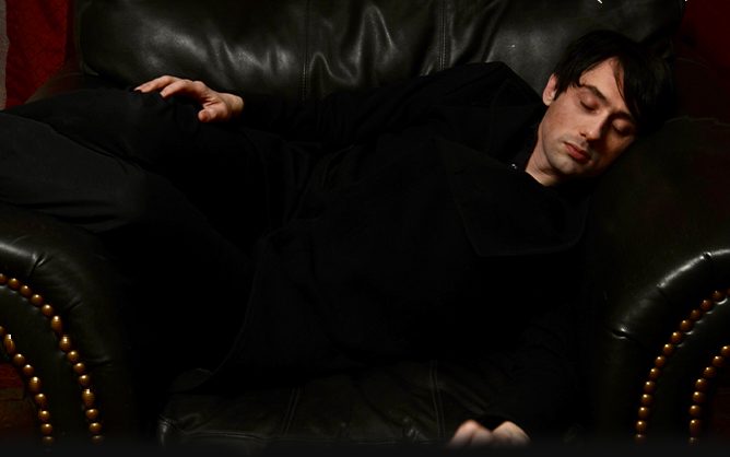 A young man, Joseph Keckler, in black lying on a couch with his eyes closed.