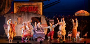 A crowd scene of 'Candide'. The dancing cast is dressed in period costume. There is a faded caravan behind them.