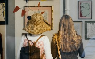 Take your date on a gallery visit for Valentine’s Day 2025. A photo of two women standing side by side with their backs to the camera, looking at a wall of drawings and a hanging mobile.