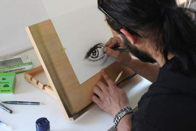 creative arts university electives: a young man pictured drawing an eye in charcoal on paper, sitting at a desk with a desk easel propping up his work.