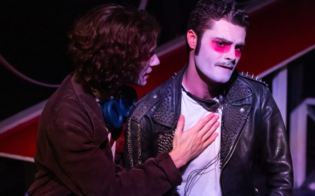 Two men. One has his hand on the chest of another, who is wearing a white t-shirt and black leather jacket. There is white make up on his face with a red streak over his eyes.