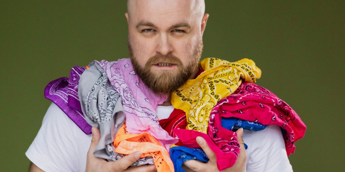 A bald man with a beard is wearing a white t-shirt. An array of different coloured scarves are drapped over his shoulder.