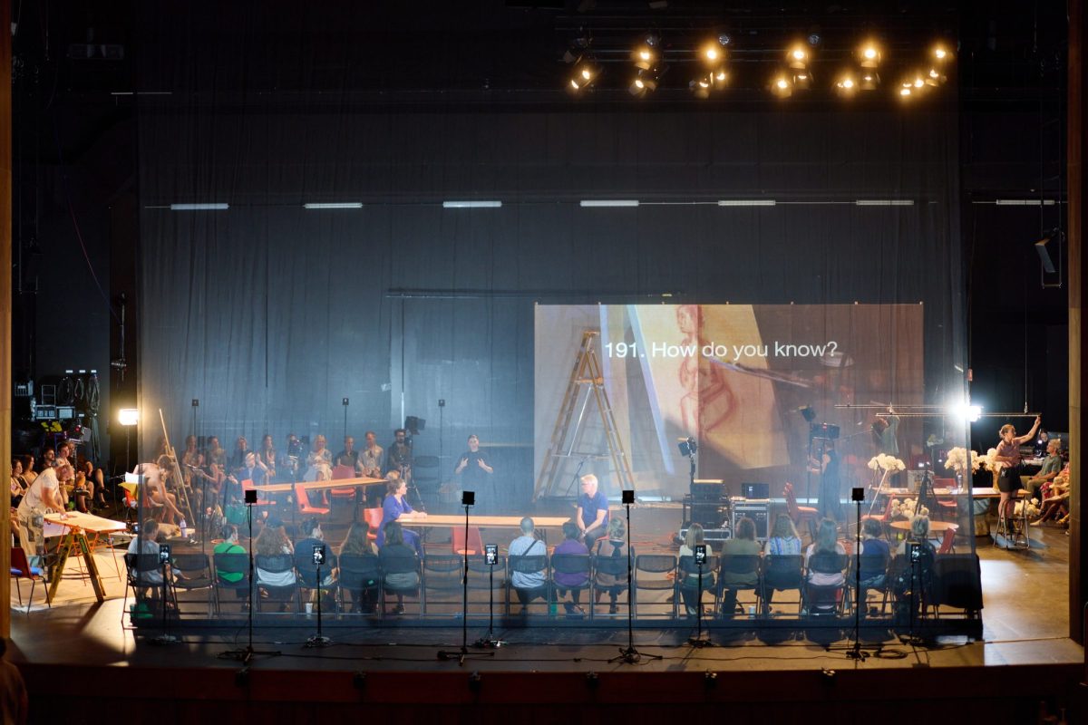 A crowd of people are seated around a central area. In the middle of which is a screen that says "191. How do you know?"