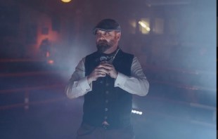 A man in a cap and a vest over a white shirt. There is mist around him.