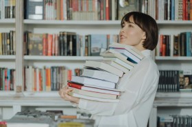 Young woman holding a stack of books with shelves in background. art books