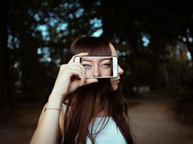 Woman with brown hair holding up phone with picture of eyes. Arts news.