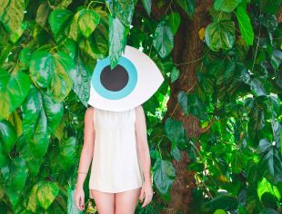 Keep your eye on this week's top arts news stories with ArtsHub's arts news watch column. Photo depicts a woman in a white sleeveless dress standing amidst the bright green leaves of a tree; her face is obscured by a giant cartoon eye.