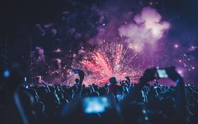 Concerts touring to Australia in second quarter of 2025. Photo of a crowd gathered outdoors, some holding up their phones to capture the fireworks in the distance.