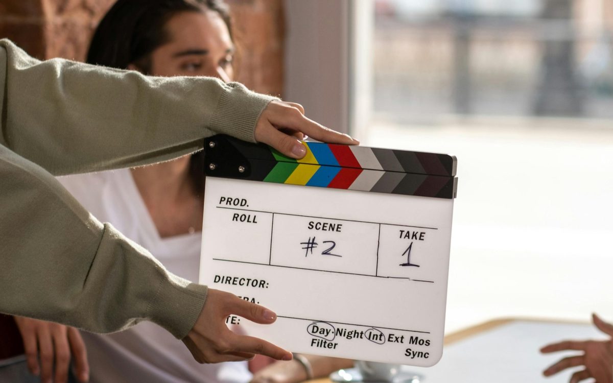 a clapperboard is centre of frame, behind are two people at a cafe table next to a window. One has only their hand seen, the other is of indeterminate gender and race. conscious casting.