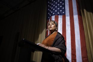 A woman standing at a podium. Behind her is an American flag.