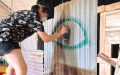 A young person wearing shorts and short sleeved shirt wearing a face mask is on stage spray painting a green eye on a corrugated iron fence.