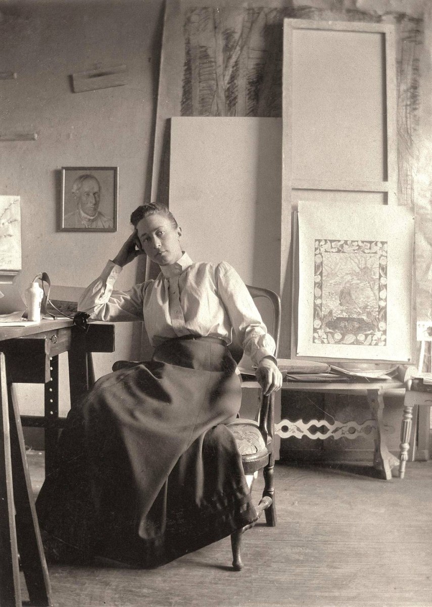 black and white image of a woman in a long skirt and white blouse sitting at a table in an artist's studio with her elbow on the table and her hand to her head. Hilma af Klint.