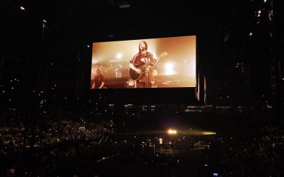 Billie Eilish 'Hit Me Hard and Soft: The Tour' in Melbourne. The singer is at the centre of a 360-degree stage holding an acoustic guitar and singing into the microphone.