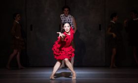 The Australian Ballet's 'Carmen', 2025, choreographed by Johan Inger, featuring Jill Ogai in the role of Carmen. A dark-haired woman in a red, ruffled dress, her legs bare, gestures dramatically and powerfully towards the camer. Three man are faintly visible in the gloom behind her. All four people are ballet dancers performing in the ballet 'Carmen'.