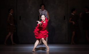 The Australian Ballet's 'Carmen', 2025, choreographed by Johan Inger, featuring Jill Ogai in the role of Carmen. A dark-haired woman in a red, ruffled dress, her legs bare, gestures dramatically and powerfully towards the camer. Three man are faintly visible in the gloom behind her. All four people are ballet dancers performing in the ballet 'Carmen'.