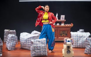 A young Cantonese Australian woman striking a funny pose with one hand behind her head and another on her waist, wearing a red Chinese-style jacket with floral patterns. Beside her is a cream-coloured robot dog with tan fluffy ears and blue glowing eyes. They are standing in front of an altar, among a loose cluster of red and blue nylon bags.