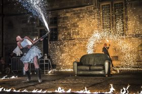 Performer holds a prop electric guitar spraying pyrotechnic sparks. In the background, a male performer waves bright sparks in a circle.