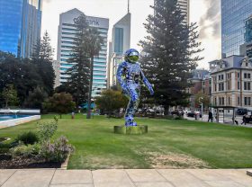 Mock-up view of 2.5-metre blue spaceman sculpture in city park surrounded by skyscrapers. Boonji Spaceman.