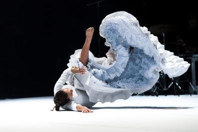 Rocío Molina, a dancing wearing a big ruffly pale blue dress. She is lying on the grown with her legs in the air that are covered by the flounces of the dress.