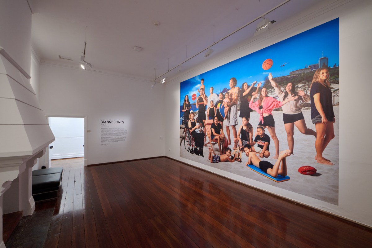 a white gallery space with a large photo on the wall of people in black and white and pink clothes playing and sitting on a beach Dianne Jones.