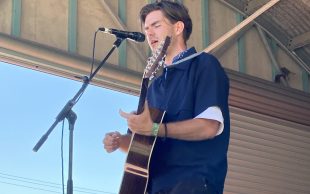 A young man in a blue shirt with an acoustic guitar plays on a small outdoor stage with his eyes closed. Flynn Gurry. Port Fairy Folk Festival.