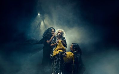 Three women. One is seated in the middle with yellow yarn. The other two gather around her.