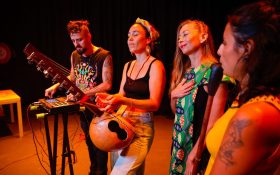 two musicians performing on stage with two other woman standing to their left. Sandra Morales is second from the right, a woman with light skin, long dark blonde hair, wearing a green patterned dress. The stage feels intimate and is cast in an orange light.