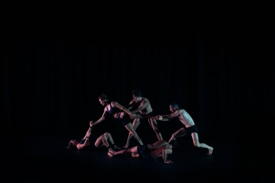 Five dancers wearing black undergarments are holding onto each other in formation.