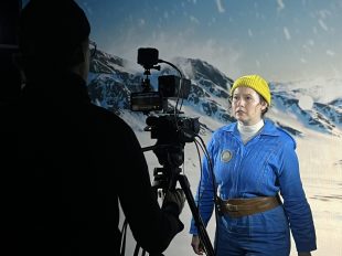 A woman in a blue jumpsuit and yellow beanie facing a video camera. The background is of snowy mountains.