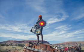 A young First Nations child wearing a mask, cape and holding a shield with Aboriginal colours, black red and yellow, standing on discarded vehicles against the Australian desert landscape.