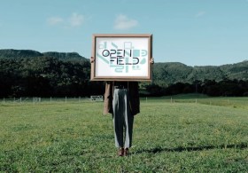 Woman standing in green landscape holding up sign over face. OpenField