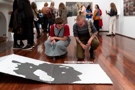 Two women looking at a printed artwork on the floor. There is a crowd behind them looking at other art.