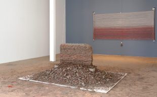 An installation work with organic material. At the centre of the space appears to be a short crumbling wall of brown, gray and slightly red debris, while on the wall behind it are rows of white and red theads outstretched with a metal frame.