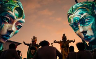 Two large installations looking like female heads decorated with face paint while two performers are dancing on the ground in front of an audience at dusk. Nuanu, Bali.