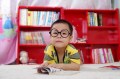 A young Asian boy with glasses and a yellow top is sitting in front of a book. There's a red bookcase behind him with books.