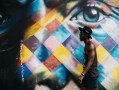 Dark skinned man wearing hat walking past colourful mural of a face with eye. Arts news watch