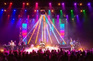 A concert scene with lots of bright lights and backdrop. Four figures in cream can be seen on stage and a back up band behind them.