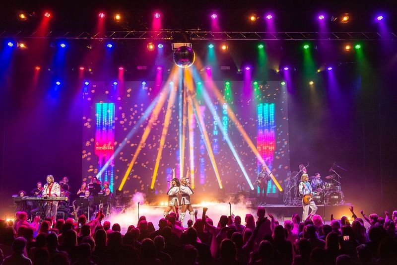 A concert scene with lots of bright lights and backdrop. Four figures in cream can be seen on stage and a back up band behind them.