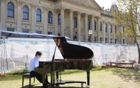 A young Asian man sitting by a grand piano outside the South Melbourne Town Hall, surrounded with scaffolding.